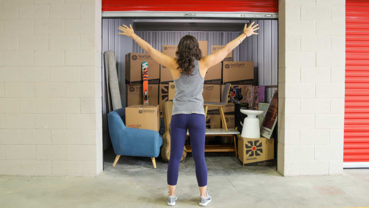 A woman standing with her arms held out in front of a storage unit filled with furniture