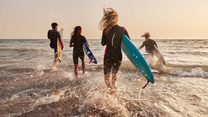 Four surfers in wetsuits clutch surfboards and are running into the ocean with the wind blowing through their hair.