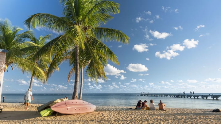 Four people sitting on a beach with a pile of kayaks behind them. Another person is walking on the beach to their left.