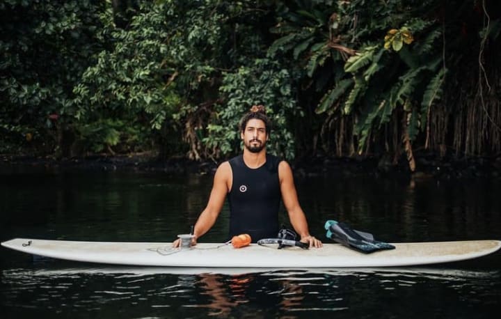 Dr. Cliff Kapono standing in hip-high water, with his hands on a surfboard that's in the foreground