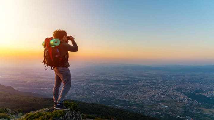 Man standing on the top of a mountain with a backpack with a yoga mat and looking through binoculars at a city below him.