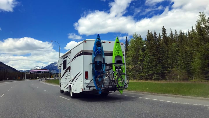 An RV with two kayaks and a bike on the back moving along a highway with mountains in the background