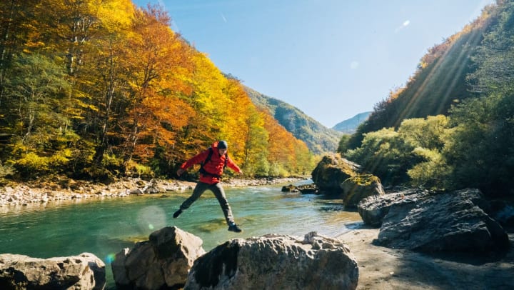 Man jumps between rocks at a river with fall colors in the background